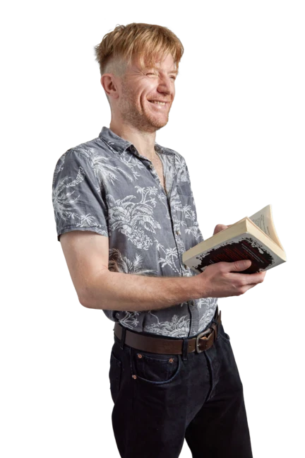 Jim is wearing a plaid shirt and black trousers as he sits on a wooden stool with his legs crossed. He holds an orange pen and looks directly into the camera, attempting to look scholarly.